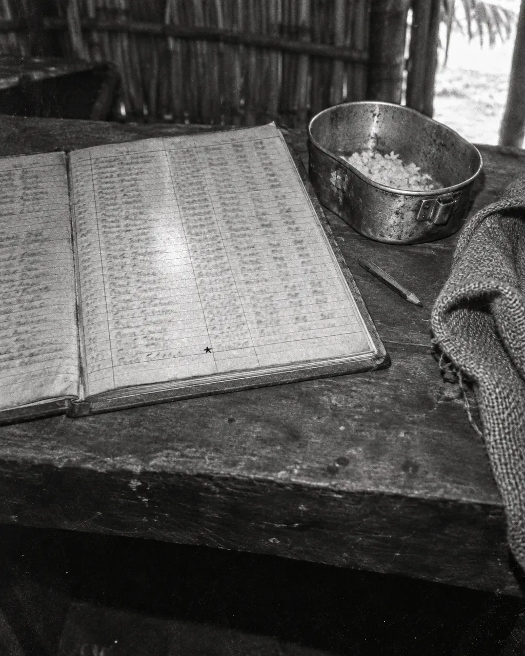 Libro de registro abierto sobre una mesa de tablones dentro de una choza con techo de palma, con columnas llenas de anotaciones manuscritas