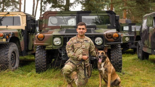 Soldado arrodillado junto a un perro malinois frente a vehículos militares