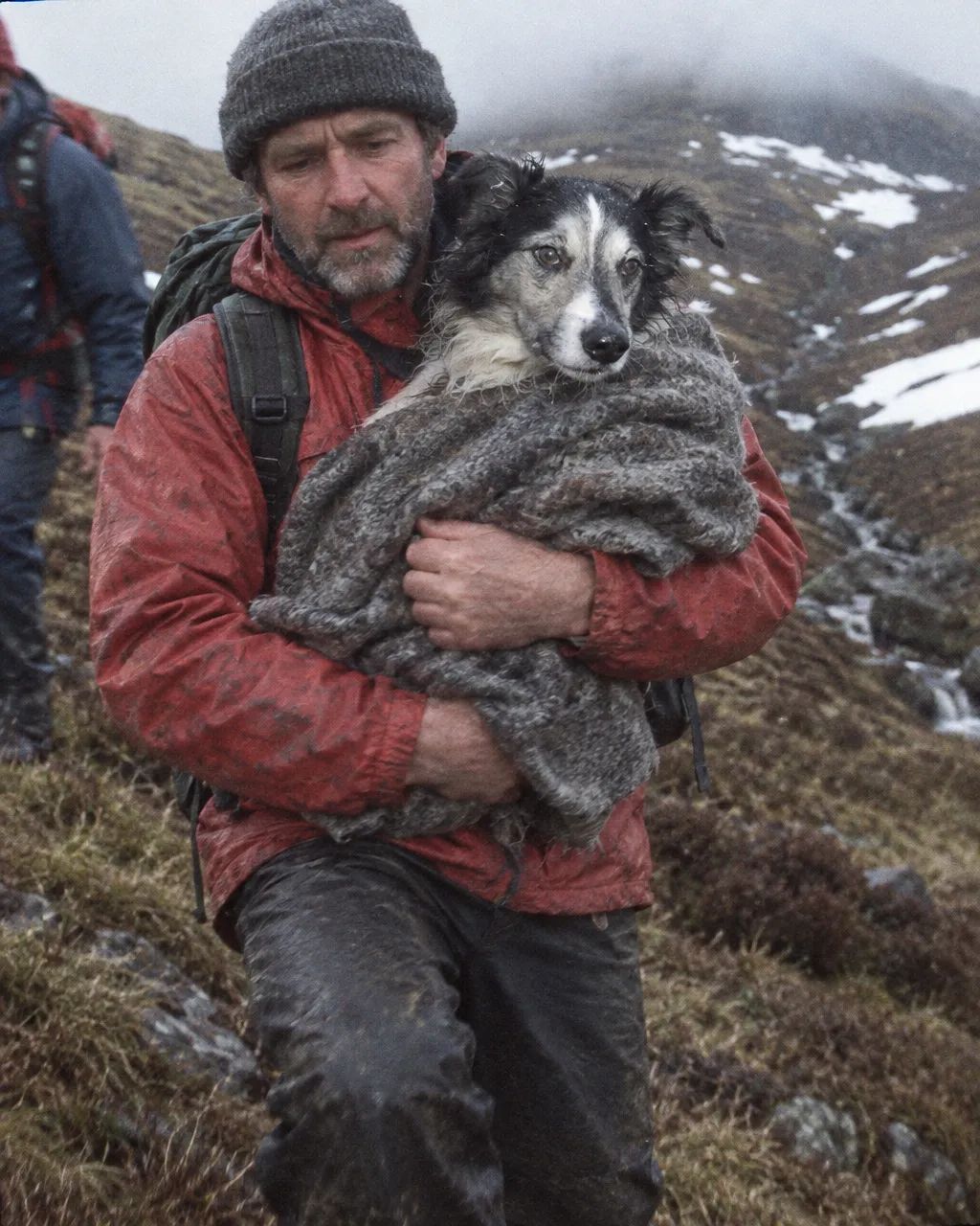 El perro que firmó con su pata para salvar un tren y luego pasó 77 noches de invierno en una montaña de Gales 5 Rescatista de montaña con chaqueta roja que baja por una ladera mojada con un perro viejo blanco y negro en brazos, envuelto en una manta gris