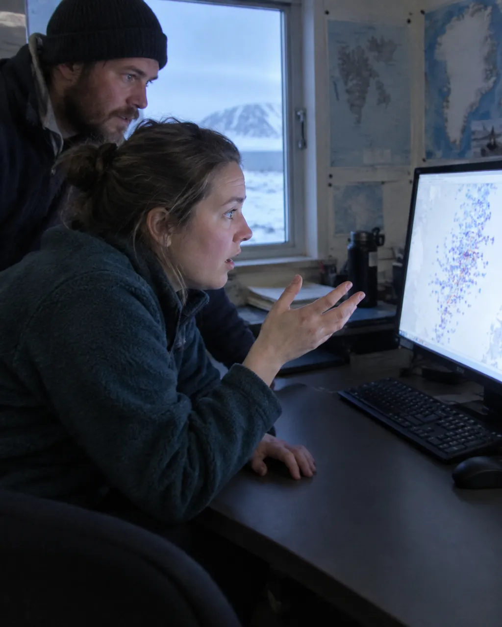Una zorra ártica de menos de un año caminaba sobre el mar helado, y nadie creyó los datos 4 En una oficina de investigación polar, una persona con chaqueta polar se inclina hacia un monitor con una mano a medio levantar, mientras otra persona mira la pantalla por encima de su hombro.